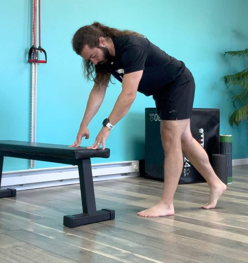 Dr. John in a split stance with his weight on his forward leg (Left) leaning over a bench with hands about to be placed on the bench as a hamstring strain healing exercise